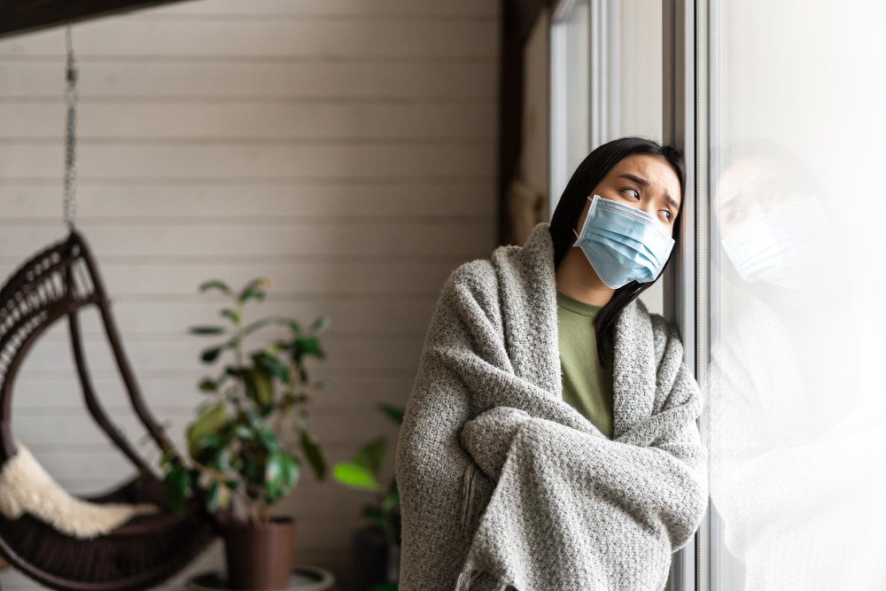 Wanita dengan masker wajah dan selimut bersender di kaca